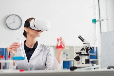 Laboratuvarda mikroskop ve test tüpleri üzerinde çalışırken elinde mataralar tutan VR kulaklık bilimcisi. 