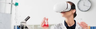 VR kulaklıklı bilim adamı mikroskop yanında mataralar ve ekipmanlar tutuyor. Laboratuvarda, afişte. 