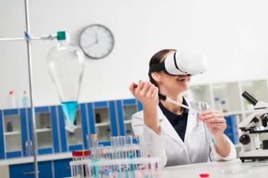 VR kulaklıklı neşeli bilim adamı laboratuarda test tüpü ve elektronik pipetle çalışıyor. 