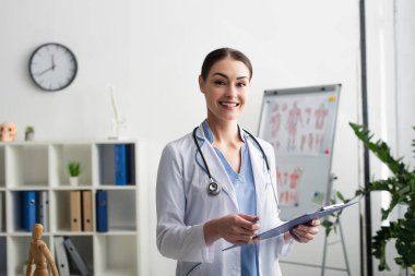 Klinikte elinde kalem ve dosya tutan neşeli esmer doktor. 