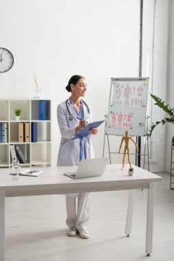 Klinikteki aletlerin, suyun ve defterin yanında gülen doktor yazı yazıyor. 