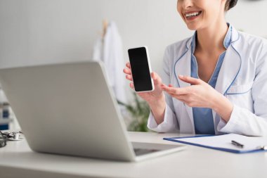 Klinikte dizüstü bilgisayardaki video konsültasyonu sırasında elinde akıllı telefon tutan gülümseyen bir doktorun görüntüsü. 