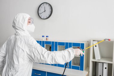 Koruyucu kıyafetli bilim adamı spreyle laboratuarı dezenfekte ediyor.