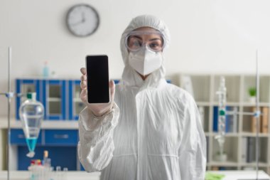 Tehlikeli madde giysisi içindeki bulanık doktor laboratuarda boş ekranla cep telefonu tutuyor.