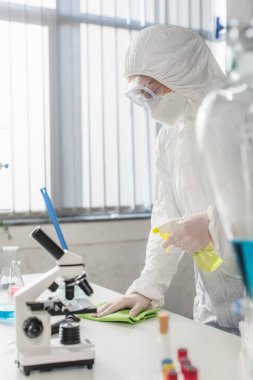 Laboratuvarda mikroskop yakınındaki koruyucu giysili doktor masasını temizliyor.