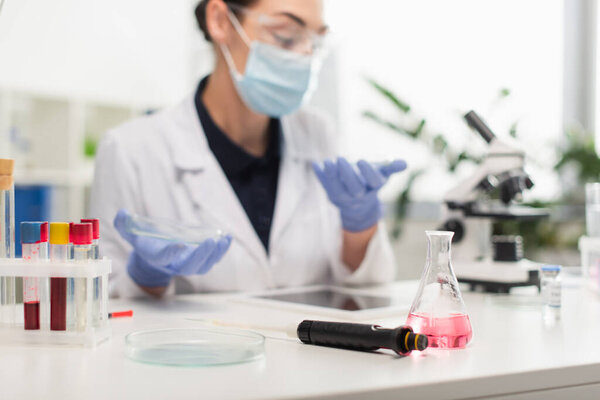 Electronic pipette, flask and petri dish near blurred scientist in laboratory 