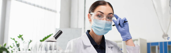 Scientist in goggles and medical mask holding pen near microscope in laboratory, banner 