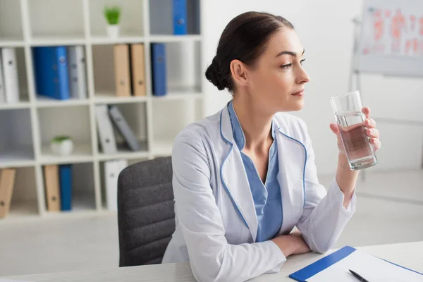 Brunette Médecin Tenant Verre Eau Près Presse Papiers Dans Clinique — Photo