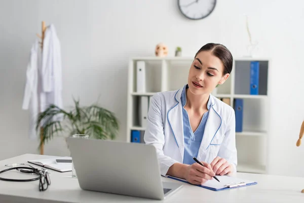 Doctor Escribiendo Portapapeles Mirando Portátil Cerca Anteojos Estetoscopio Clínica —  Fotos de Stock