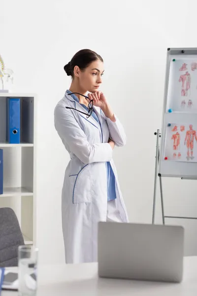 Médico Pensativo Segurando Óculos Enquanto Estava Perto Laptop Borrado Flipchart — Fotografia de Stock