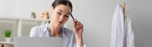 Przemyślany Lekarz Trzymający Długopis Patrząc Niewyraźny Laptop Szpitalu Baner — Zdjęcie stockowe