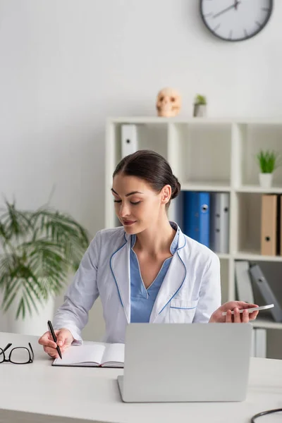 Morena Médico Sosteniendo Teléfono Inteligente Escritura Portátil Cerca Borrosa Portátil — Foto de Stock