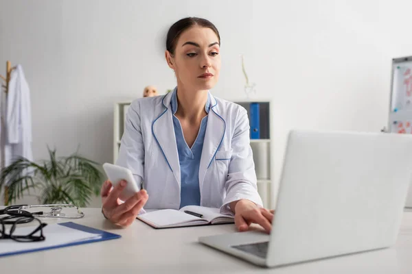 Brunetka Lekarz Trzymając Telefon Komórkowy Podczas Pisania Zamazany Laptop Klinice — Zdjęcie stockowe