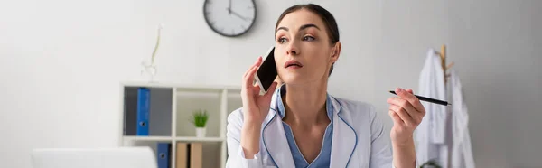 Médico Segurando Caneta Olhando Para Longe Enquanto Fala Smartphone Clínica — Fotografia de Stock