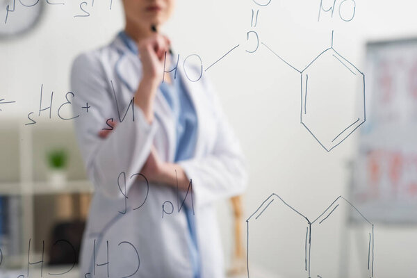 partial view of blurred doctor standing near glass board with chemical formulas
