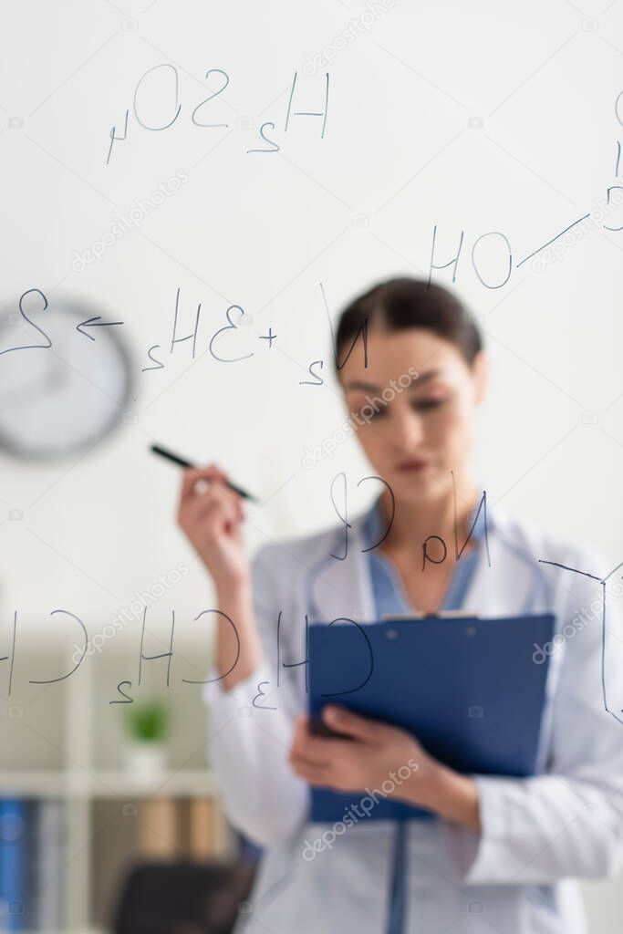 selective focus of chemical formulas on glass board near scientist with clipboard and pen