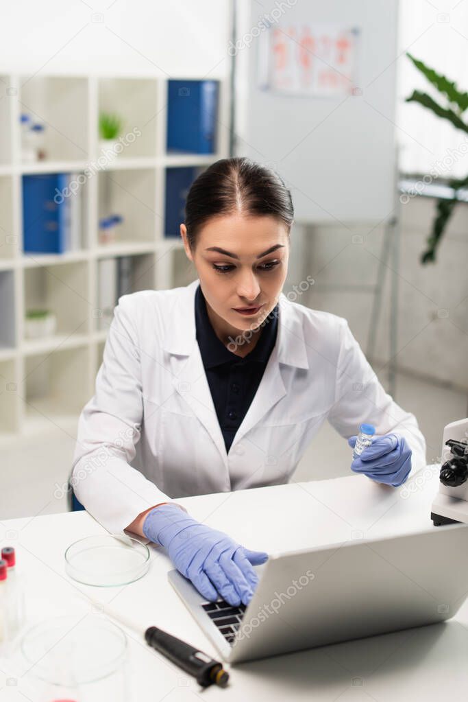 brunette scientist holding vaccine while typing on laptop near petri dish