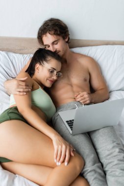 Üstsüz bir adam yatağında dizüstü bilgisayarın yanında seksi bir kadına sarılıyor. 