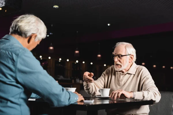 Пожилой Мужчина Очках Разговаривает Размытым Другом Возле Чашек Кофе — стоковое фото