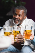 senior africký Američan muž s úsměvem při cinkání rozmazané pivo brýle s přáteli