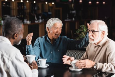Kafede çok ırklı arkadaşlarla konuşurken neşeli Asyalı adam el kol hareketi yapıyor.