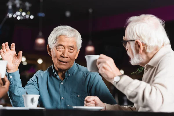 senior asian man gesturing while drinking coffee and talking with multiethnic friends