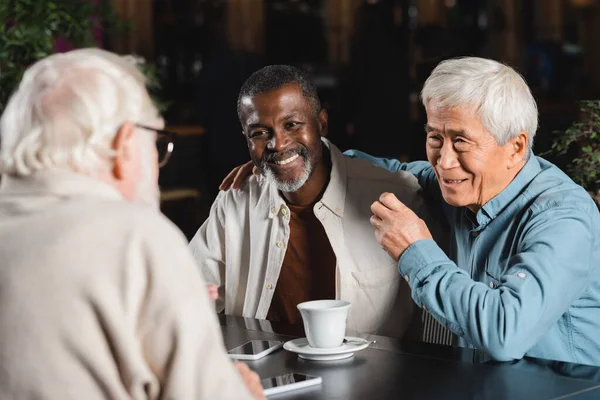 smiling asian man embracing shoulders of african american friend in cafe