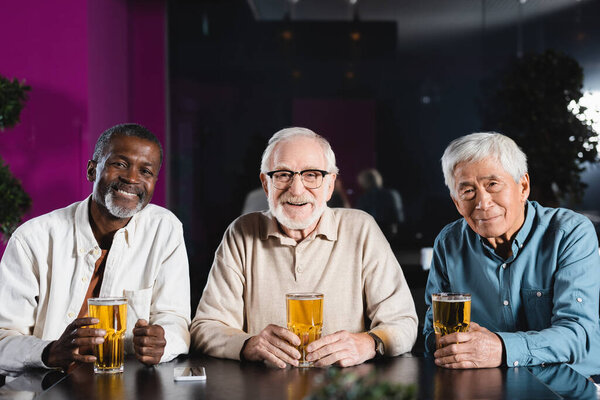 счастливые старшие многонациональные друзья, смотрящие в камеру, сидя в пабе со стаканами пива