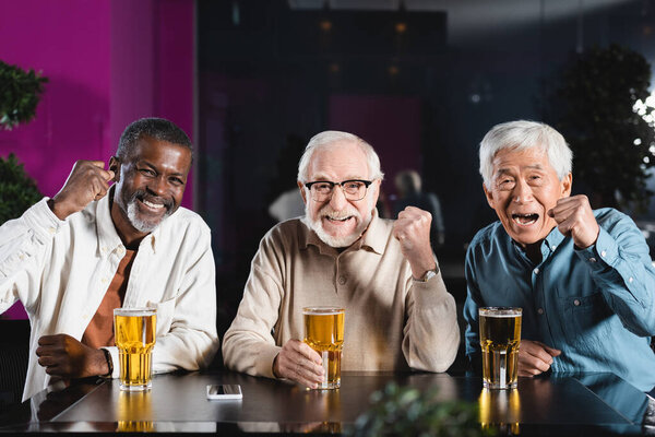 счастливые старшие многонациональные друзья, показывающие победный жест во время просмотра чемпионата по футболу в пабе