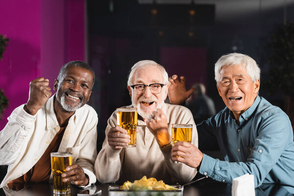 веселые старшие мультикультурные друзья с бокалами пива показывают победу жест во время просмотра футбольного матча в пабе
