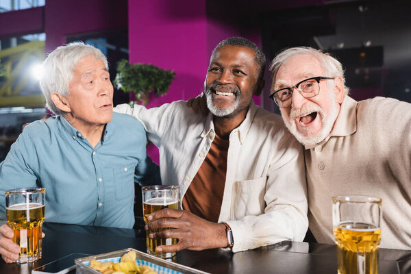 cheerful elderly multicultural friends watching football championship in beer pub