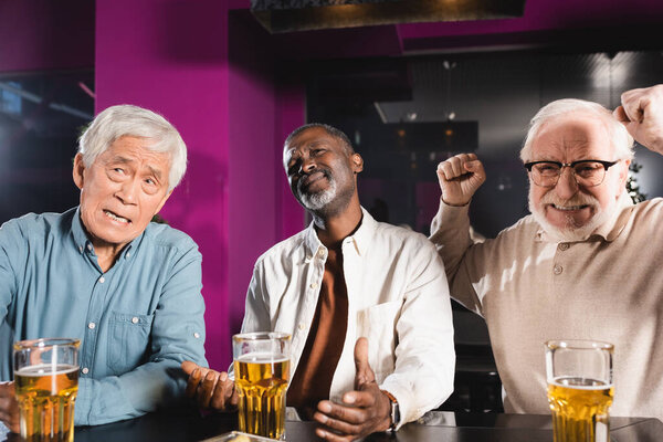 взволнованный пожилой человек показывает жест успеха во время просмотра футбольного матча с расстроенными межрасовыми друзьями