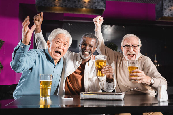 взволнованные пожилые многонациональные друзья, показывающие жест триумфа во время просмотра чемпионата по футболу в пабе