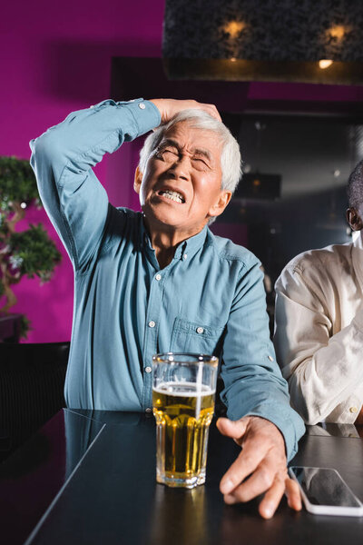 shocked senior asian man with closed eyes touching head while sitting with african american friend in sports bar