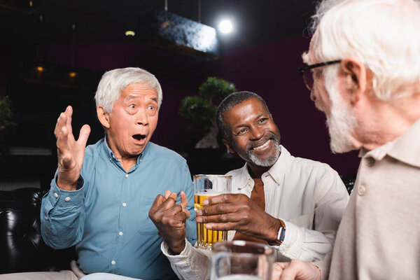 astonished asian man gesturing while talking to elderly multiethnic friends in beer pub