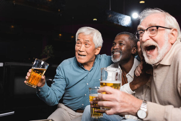 изумленный пожилой человек с открытым ртом смотрит футбол в пивном пабе со счастливыми межрасовыми друзьями