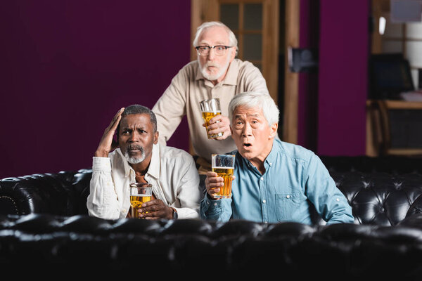 шокированные старшие многонациональные друзья с бокалами пива, смотрящие футбольный чемпионат в пабе