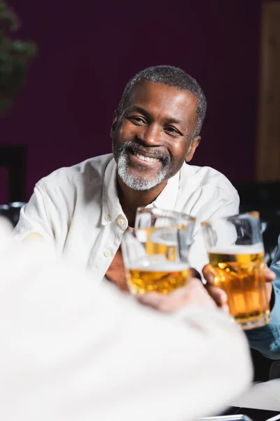 Wesoły Afroamerykanin Mężczyzna Brzęczy Szklanki Piwa Zamazanymi Starszymi Przyjaciółmi Pubie — Zdjęcie stockowe