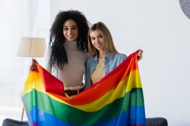 Genç ve neşeli çok ırklı lezbiyenler ellerinde lgbt bayrağıyla kameraya bakıyor.