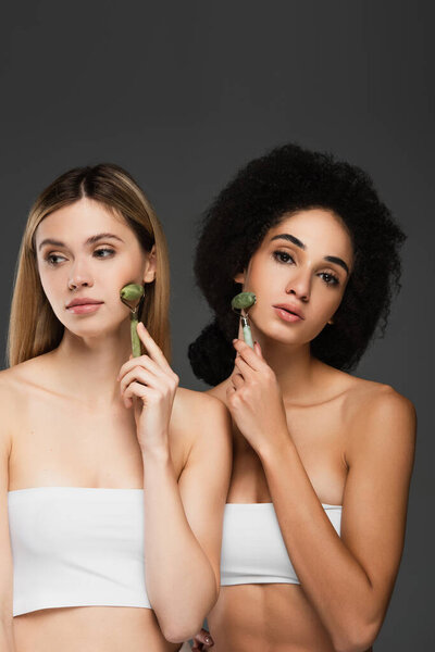 young multiethnic women massaging faces with jade rollers isolated on grey