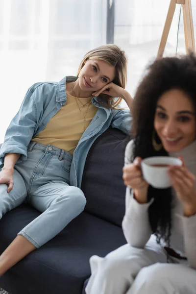 快乐的女同志看着模糊的非洲裔美国女友在沙发上喝茶 — 图库照片