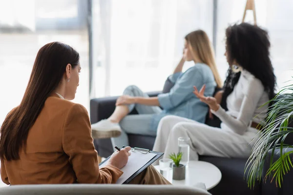 Psycholoog Schrijven Klembord Buurt Van Interraciale Lesbische Paar Ruziën Bank — Stockfoto