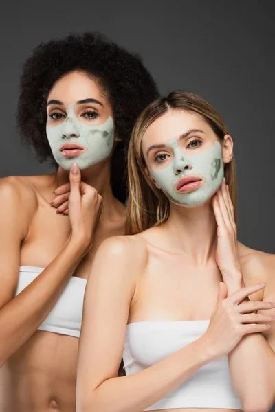 pretty multiethnic women in clay masks looking at camera while posing isolated on grey