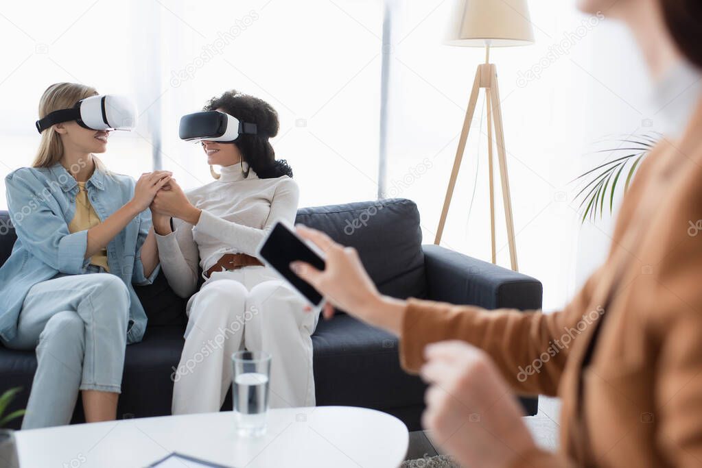 smiling same sex couple in vr headsets holding hands on sofa near blurred psychologist with smartphone 