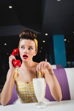 Şaşırmış bir kadın klasik bir telefonla konuşuyor. Bir bardak milkshake 'in yanında.
