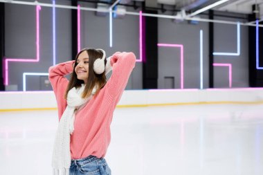 Pembe kazaklı, eşarplı ve buz pistinde kulaklık takan gülümseyen genç kadın. 