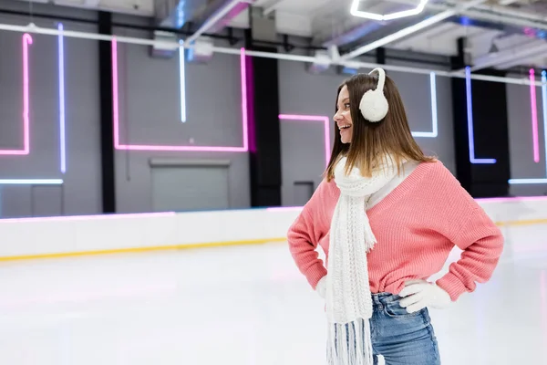 happy woman in ear muffs, scarf and pink sweater standing with hands on hips on ice rink