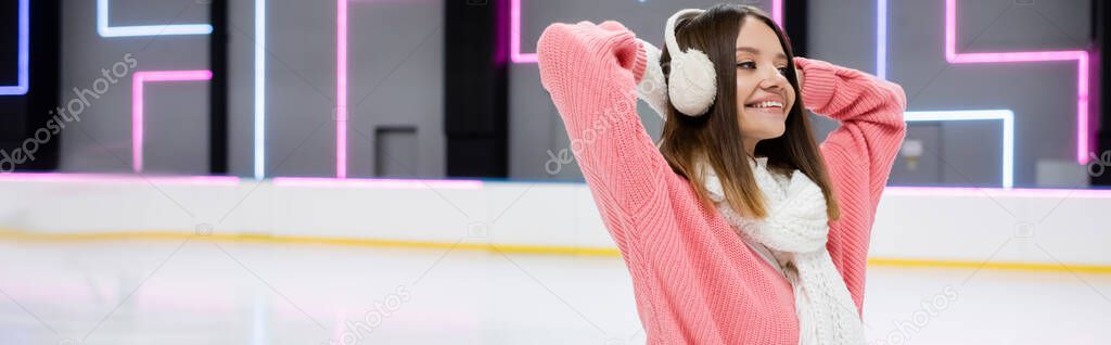happy young woman in ear muffs and scarf on ice rink, banner