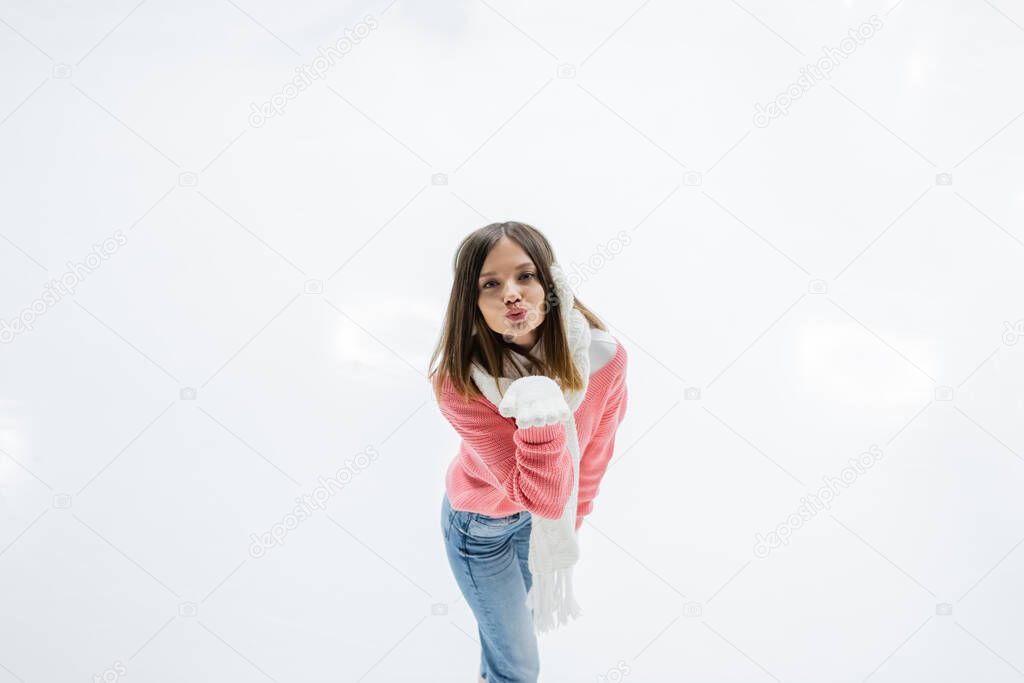 high angle view of young woman in ear muffs and scarf sending air kiss while skating on ice rink