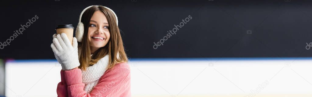 cheerful young woman in ear muffs and scarf holding paper cup on ice rink, banner